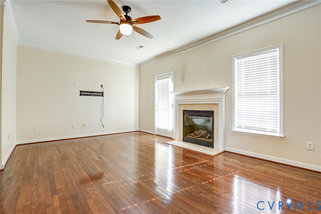 Living area featuring wood-finish flooring, a traditional fireplace with a white mantel, crown molding, and a ceiling fan