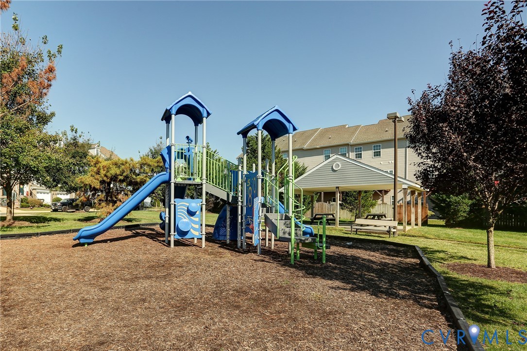 Playground structure with multi-level platforms, dual slides, and climbing elements set within a wood-mulched area, accompanied by a covered pavilion with integrated bench seating