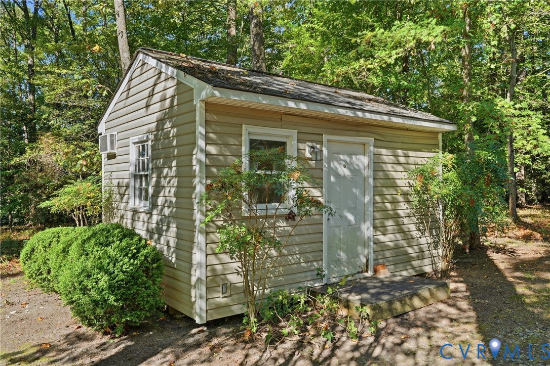 Outbuilding featuring vinyl siding, shingle roof, white trim, single-entry door, and installed wall-mounted cooling unit