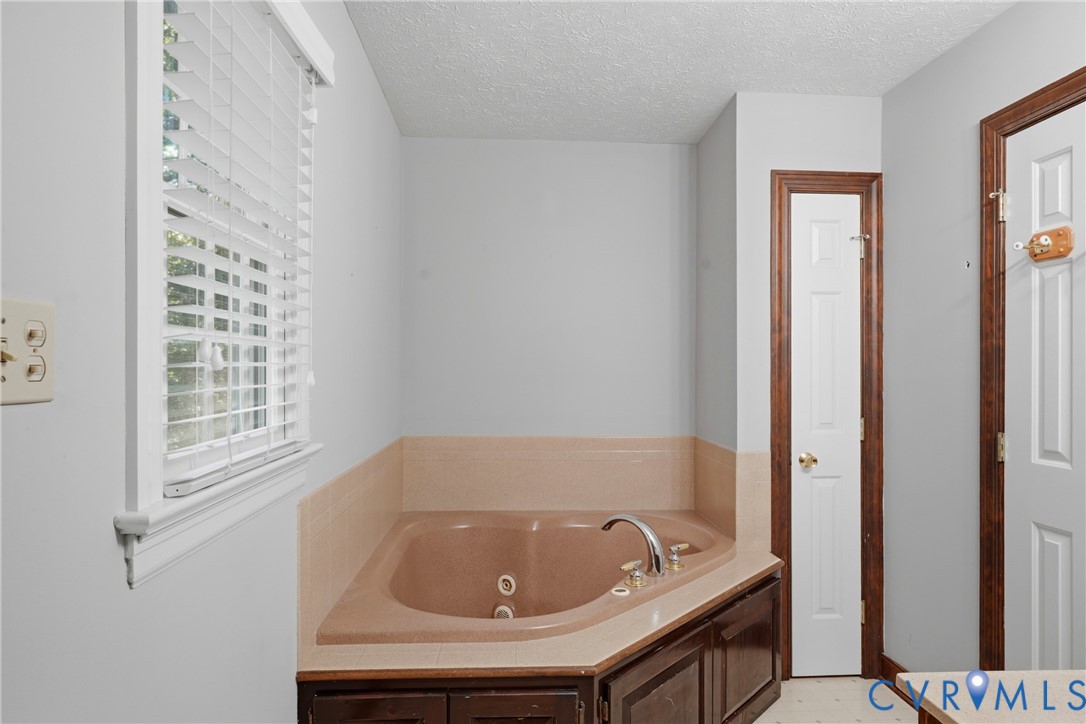 Corner jetted tub featuring a tile surround, dark wood-finish cabinetry, metal faucet fixtures, and horizontal slat window blinds