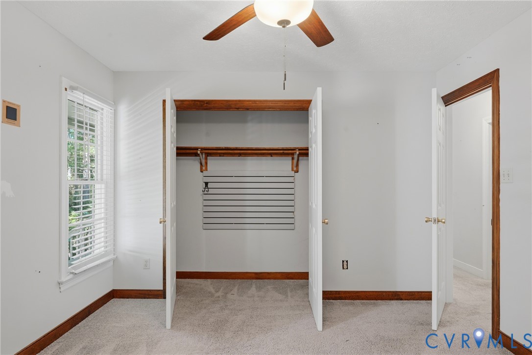 Carpeted room featuring a ceiling fan with integrated lighting, wood-trim windows, double-door closet with built-in shelving and slat wall organization, and solid wood door casings