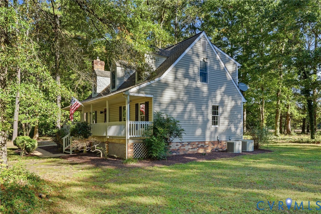 Two-story residence featuring light-colored horizontal siding, a covered wrap-around porch with railing, dormer windows, a brick foundation base, and a sprawling lot with mature trees