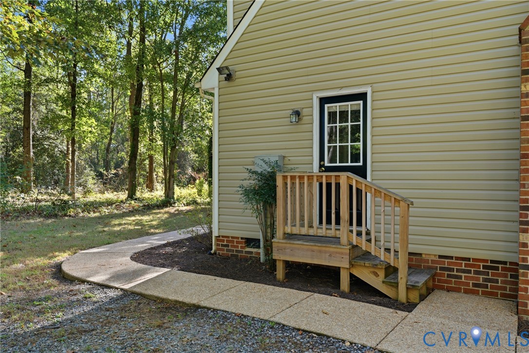 Exterior side entry with wood steps and railing, light-colored horizontal siding, brick foundation detail, and paved concrete walkway