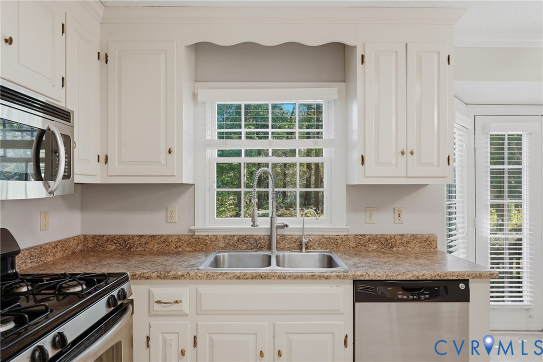 White shaker-style cabinetry with overhead scalloped detail, stone-patterned countertops, stainless steel dual-basin sink with arched faucet, and integrated stainless steel appliances