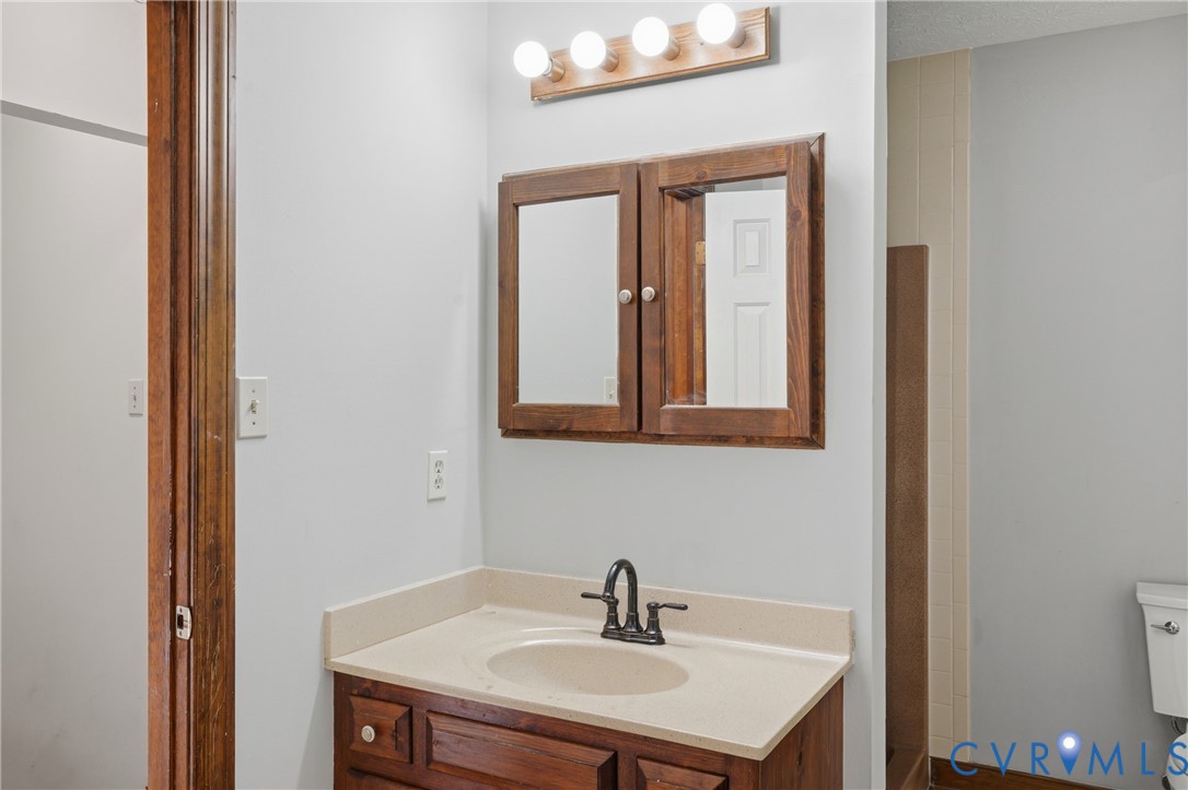 Wood-finish vanity cabinet, solid-surface countertop with integrated basin, dark metal widespread faucet, wood-framed medicine cabinet, and four-light vanity fixture