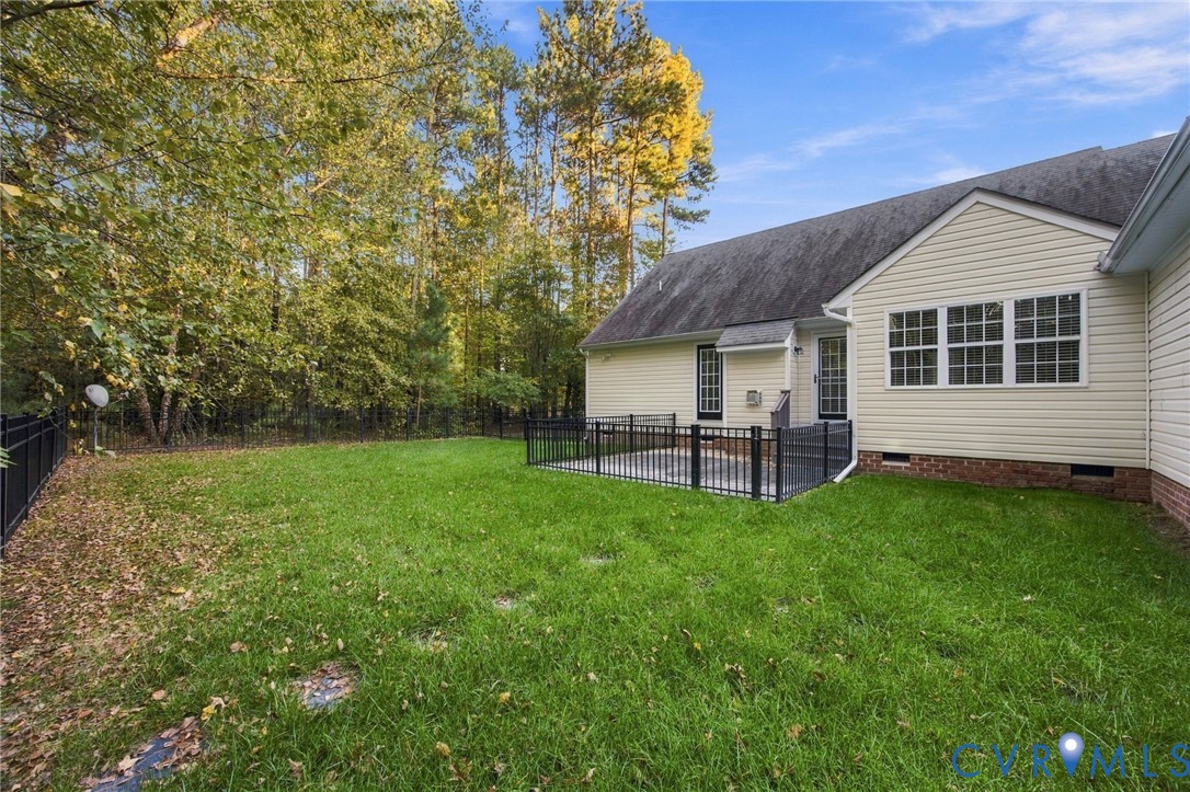 Rear exterior featuring light-toned siding, a concrete patio enclosed by metal fencing, and an expansive lawn bordered by mature trees