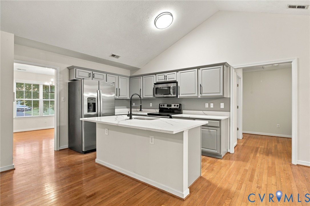 Kitchen featuring vaulted ceilings, quartz countertops, gray shaker-style cabinetry, stainless steel appliances, and wood-finish flooring
