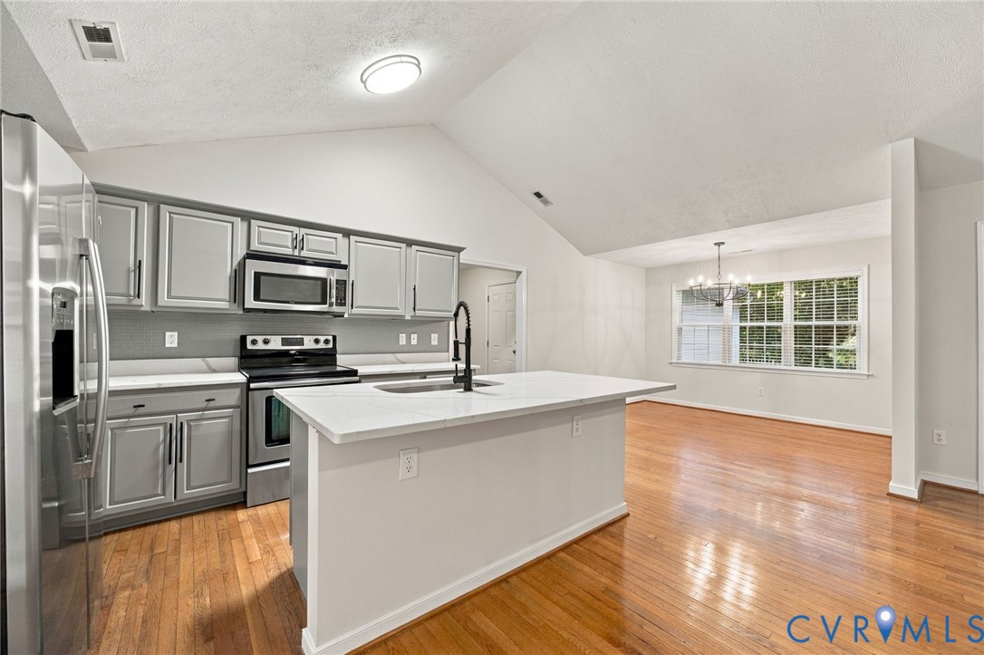 Kitchen featuring grey shaker-style cabinetry, quartz countertops, a central island with a matte black faucet, vaulted ceilings, and wood-finish flooring