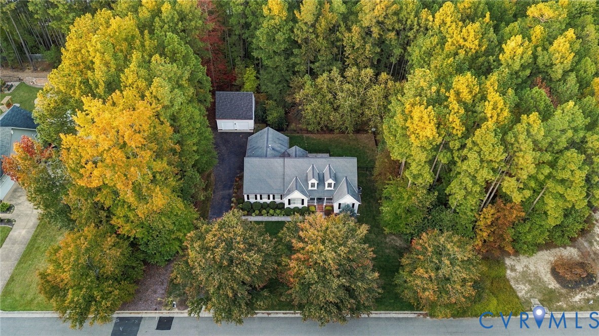 Aerial perspective of a detached residence featuring a multi-gable roofline, a detached garage, and an asphalt driveway, situated on a lot with mature deciduous trees