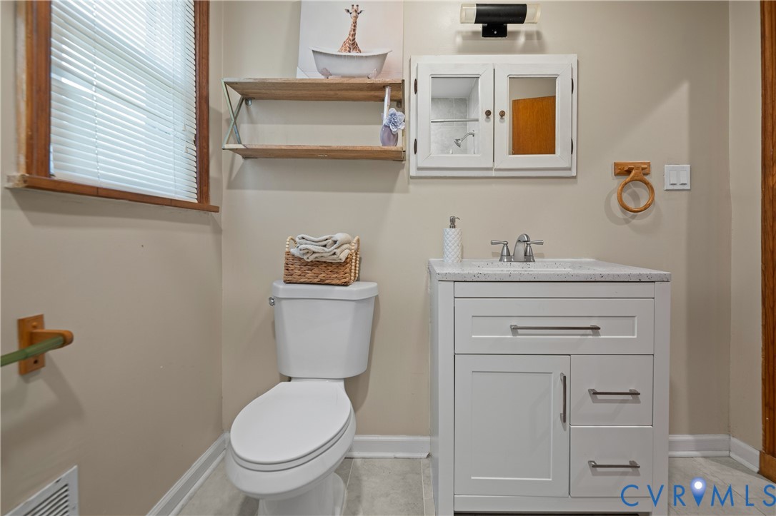 White vanity with stone-composite countertop, dual-handle faucet, surface-mount medicine cabinet, wall-mounted shelving, and tile flooring