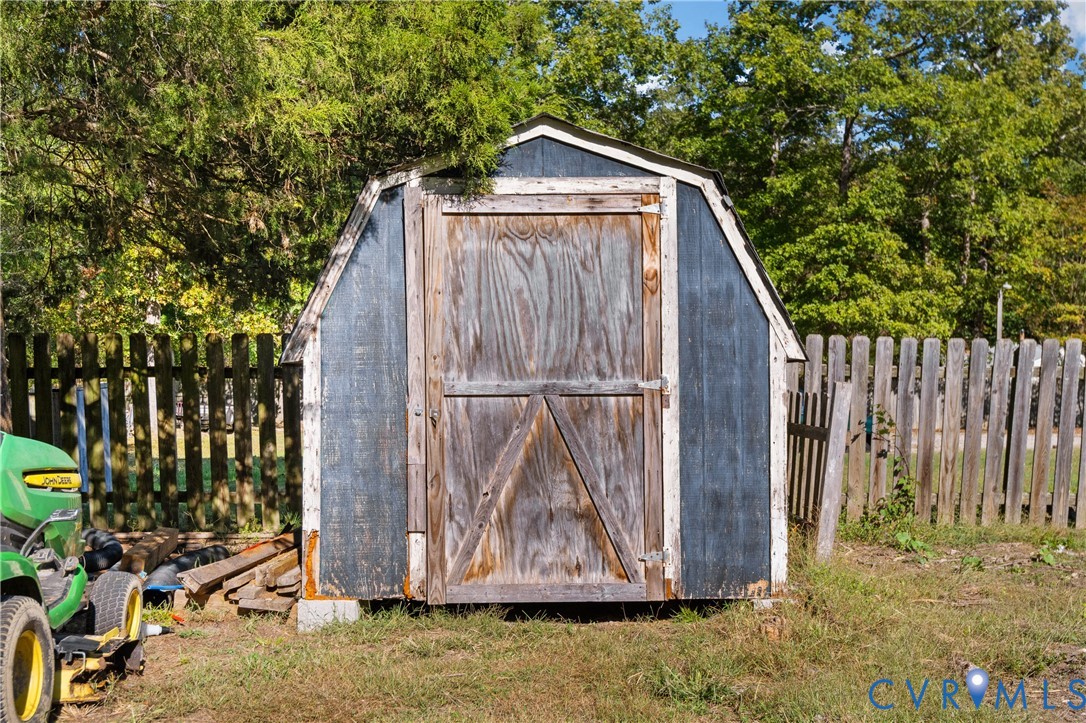 Gambrel-roof storage shed featuring wooden siding, a hinged door, and a perimeter wooden privacy fence