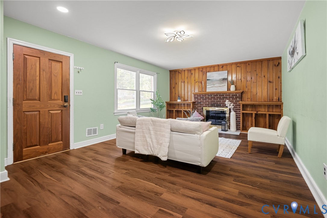 Living area featuring wood-finish flooring, wood-paneled walls, a brick fireplace, built-in shelving, and a solid wood entry door Living area featuring wood-finish flooring, wood-paneled walls, a brick fireplace, built-in shelving, and a solid wood entry door