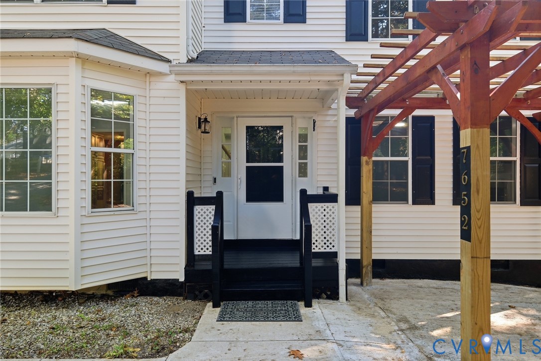 Exterior entrance featuring white siding, black shutters, a covered porch with lattice-accented railings, and a wood-frame pergola