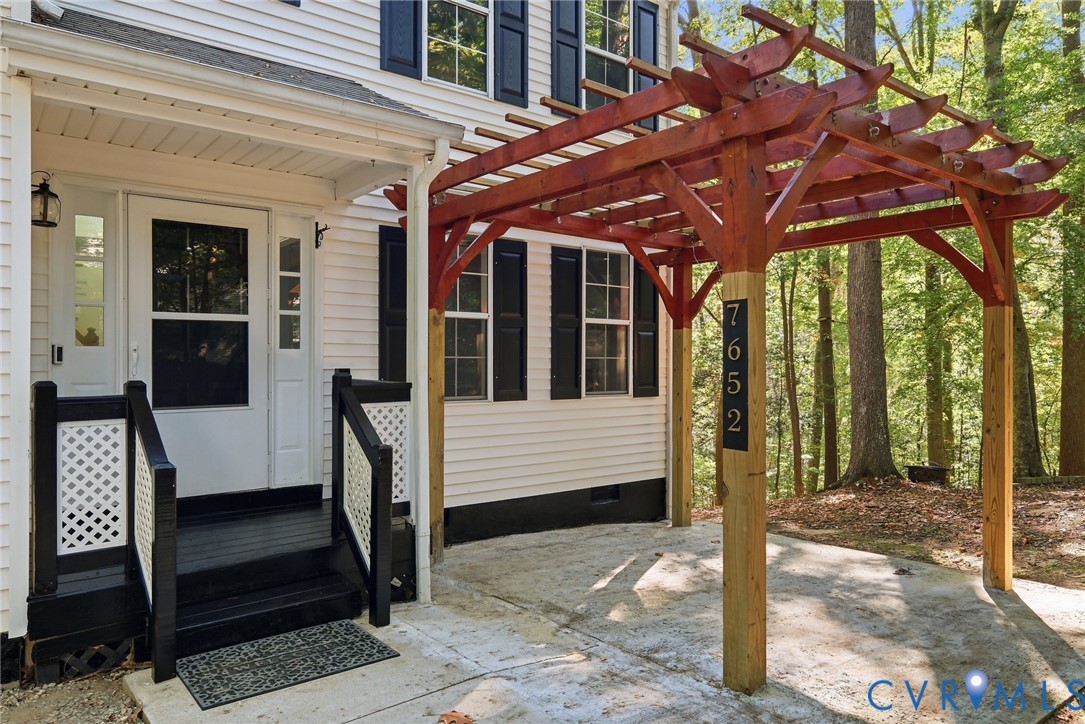 Exterior entry featuring a wood pergola, concrete patio, white lap siding, black shutters, and stairs with decorative lattice railings