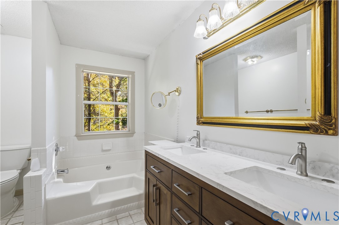 Dual vanity with stone-finish countertop, undermount sinks, wood-finish cabinetry, built-in soaking tub, and wall-mounted articulating mirror