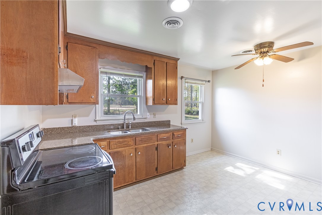 Kitchen featuring wood cabinetry, stone-surface countertops, dual-basin stainless steel sink, black electric range, and ceiling fan with integrated lighting