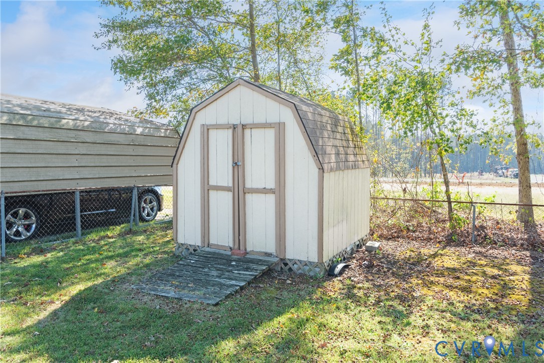 Gambrel-style storage shed with double-door entry, wood-paneled siding, and shingled roof, set on a grass-covered lot with chain-link perimeter fencing