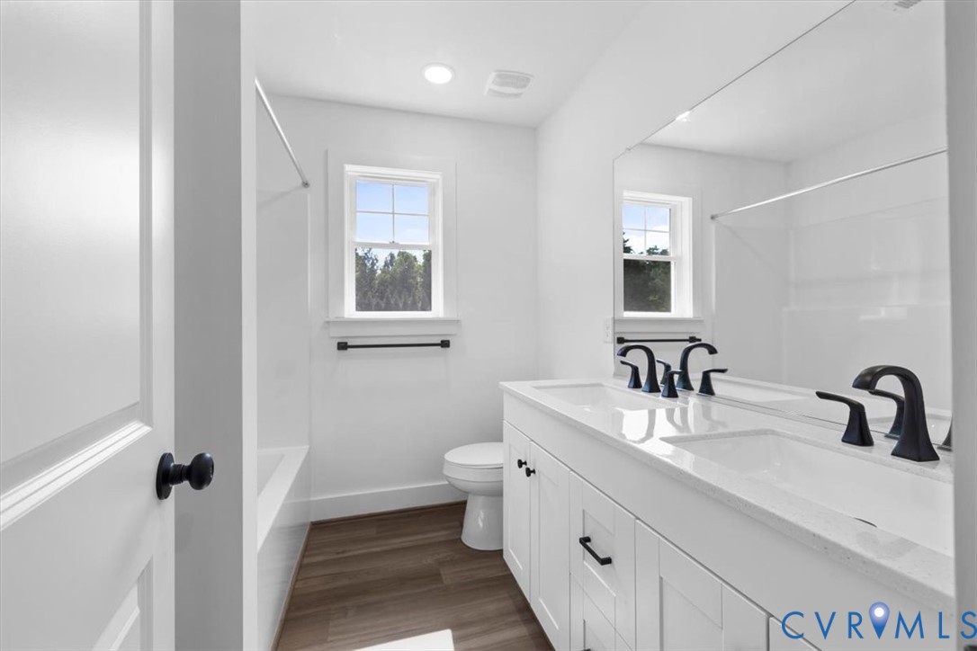Bathroom featuring a white double-sink vanity with a solid-surface countertop, matte black fixtures, recessed lighting, and wood-finish flooring
