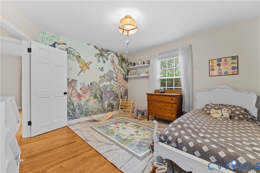 Bedroom featuring wood-finish flooring, a wall-mounted floating display shelf, a single-hung window with shutters, and a multi-panel interior door