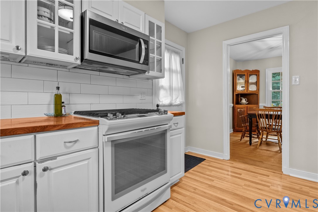 Kitchen featuring white cabinetry with glass-panel inserts, butcher-block countertops, a white subway-tile backsplash, stainless steel over-the-range microwave, and wood-finish flooring