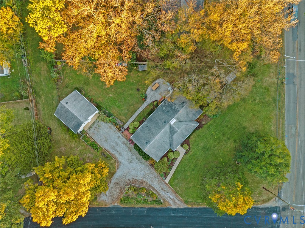 Aerial perspective of a property featuring a detached garage, expansive lawn, mature trees, gravel driveway, and paved patio area