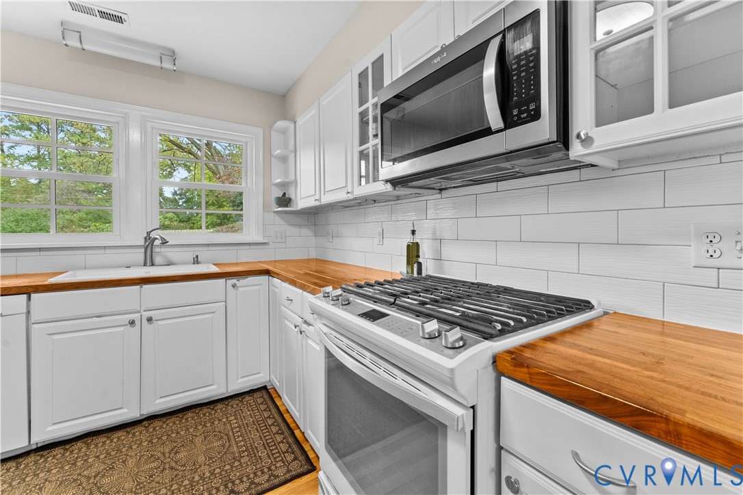 Kitchen featuring white cabinetry with glass-front uppers, wood-finish countertops, subway tile backsplash, and stainless steel appliances