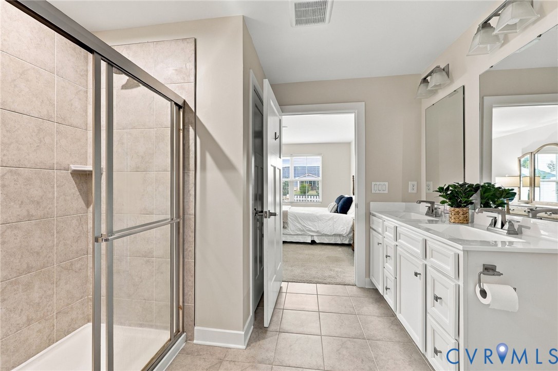 Bathroom featuring a double-sink vanity with white cabinetry, neutral tile flooring, a glass-enclosed shower with tiled walls, and dual wall-mounted light fixtures