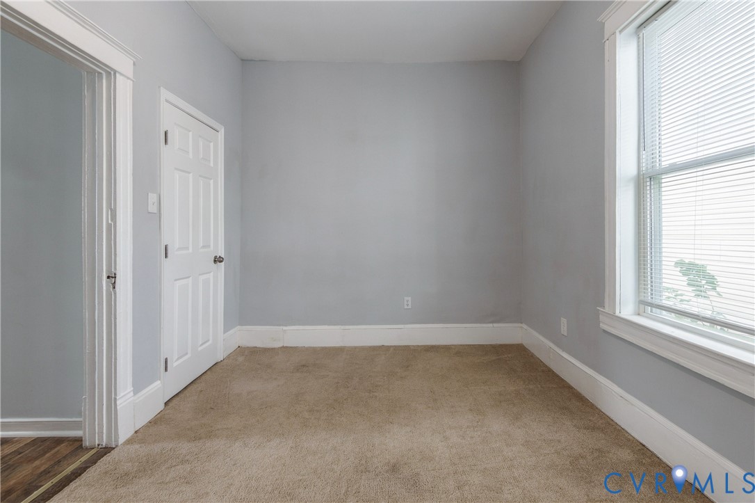 Neutral-toned interior space featuring light-colored wall paint, plush carpeting, a single-hung window with horizontal blinds, and a traditional six-panel interior door