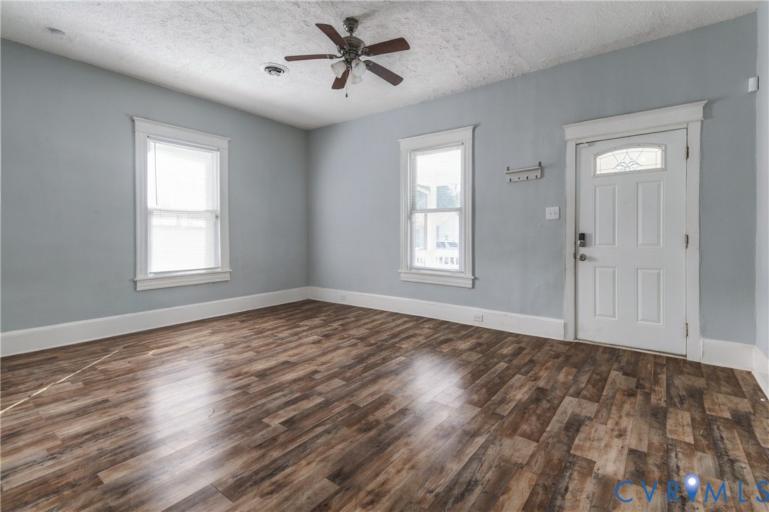 Room featuring wood-finish flooring, two windows with white trim, a multi-pane entry door with glass transom, and a central ceiling fan with integrated lighting
