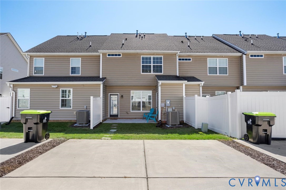 Rear exterior featuring tan siding, a dark shingle roof, white privacy partial fencing, and a concrete patio and private paved 2-car driveway.