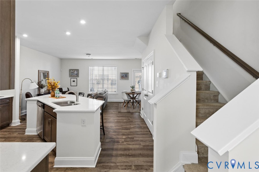 Open-concept living space with LVP flooring, a kitchen island with integrated sink, recessed lighting, and a prominent staircase with carpeting.