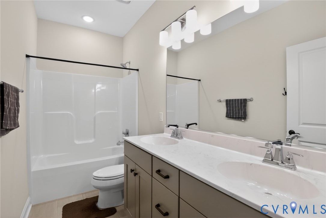 Bathroom featuring a dual-sink vanity with light-toned stone-surface countertop, white tub-and-shower combination, large wall-mounted mirror, and multi-bulb overhead lighting fixture