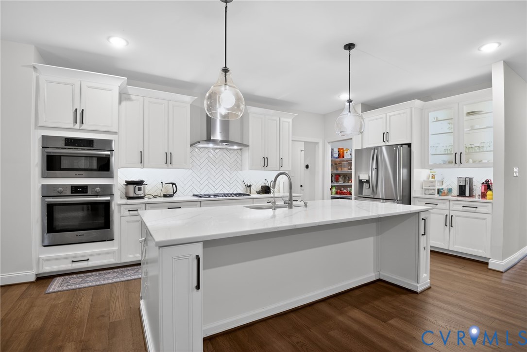 Kitchen featuring white shaker-style cabinetry, quartz countertops, a herringbone tile backsplash, stainless steel built-in appliances, and a central island with pendant lighting