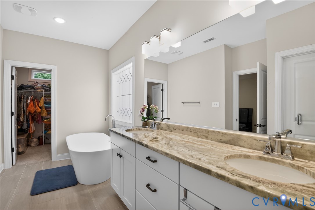 Dual-sink vanity with stone countertop and matte black hardware, freestanding soaking tub, large wall-mounted mirror, and tile flooring