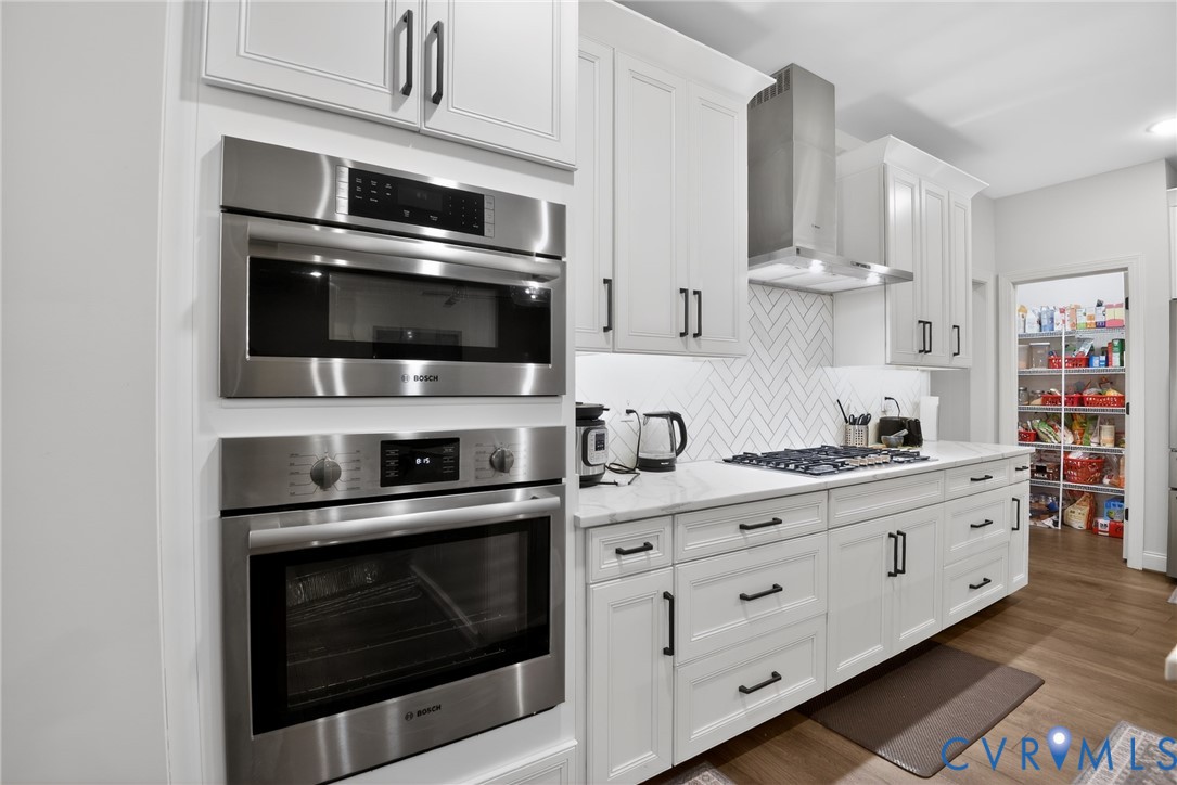 White cabinetry with matte black hardware, stainless steel built-in wall oven and microwave, stone-finish countertops, herringbone-patterned tile backsplash, and stainless steel range hood