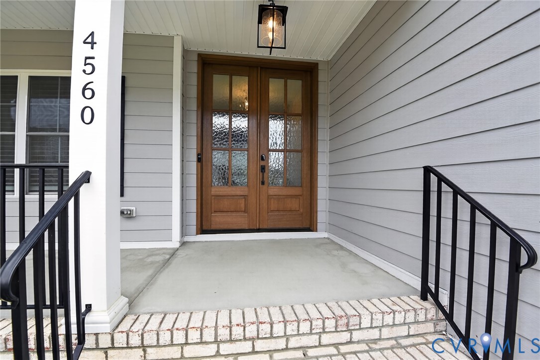 Covered front porch featuring wood-finish double entry doors with glass panels, light-gray horizontal siding, brick-base steps, and a black metal handrail