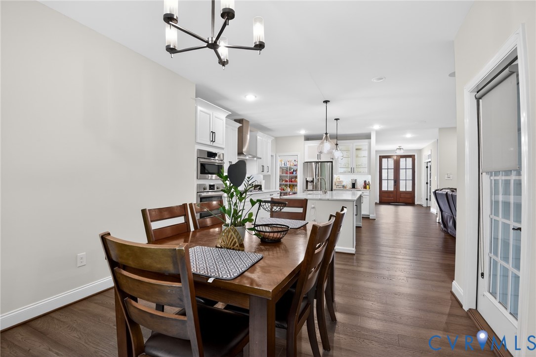 Open-concept kitchen and dining area featuring wood-finish flooring, white shaker cabinetry, stainless steel built-in double wall ovens, a kitchen island with stone countertops, and multi-light pendant chandeliers