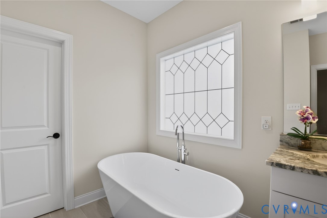 Freestanding oval soaking tub with floor-mounted chrome faucet, decorative lattice-pattern window, solid surface vanity countertop, and shaker-style cabinetry