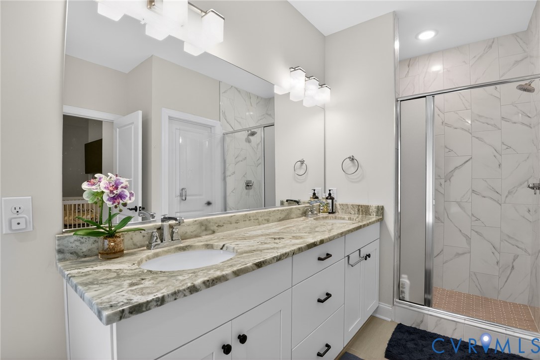Dual-sink vanity with stone countertop, white shaker-style cabinetry, matte black hardware, and floor-to-ceiling tile shower enclosure