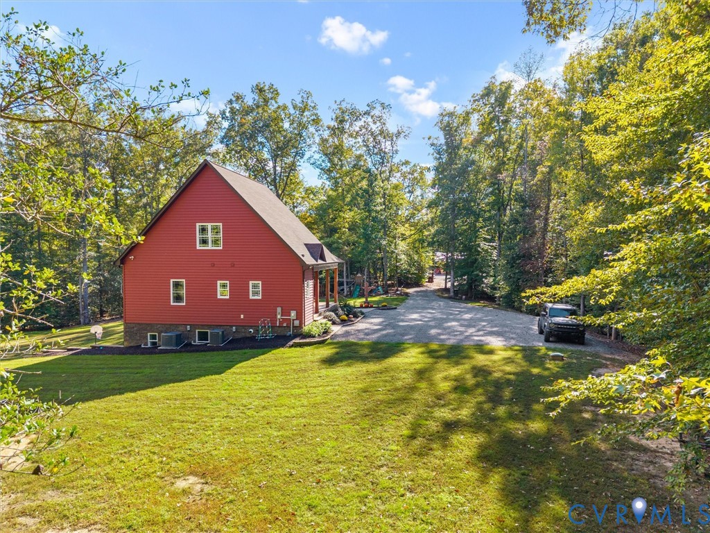 A picturesque side view of the home showcases the expansive green lawn, mature shade trees, and beautifully maintained landscaping. The spacious gravel parking area and peaceful wooded surroundings add to the charm and convenience of this country retreat!