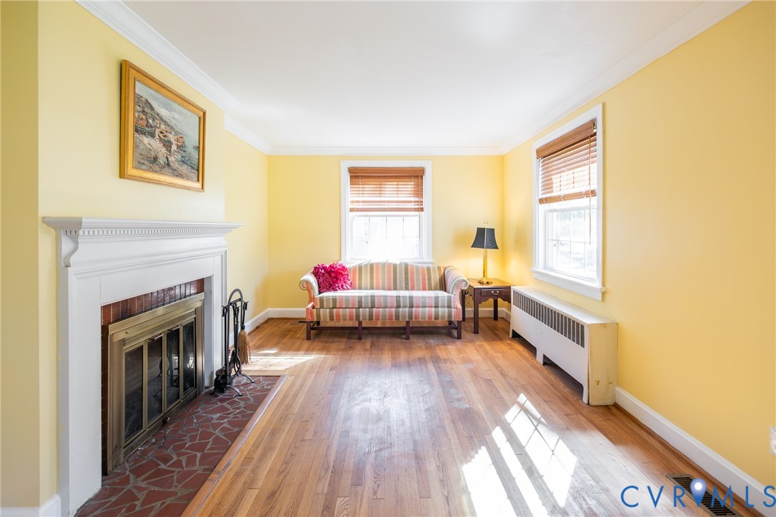 Wood-finish flooring, fireplace with a white mantel and brick hearth, crown molding, and two windows with wood blinds Wood-finish flooring, fireplace with a white mantel and brick hearth, crown molding, and two windows with wood blinds