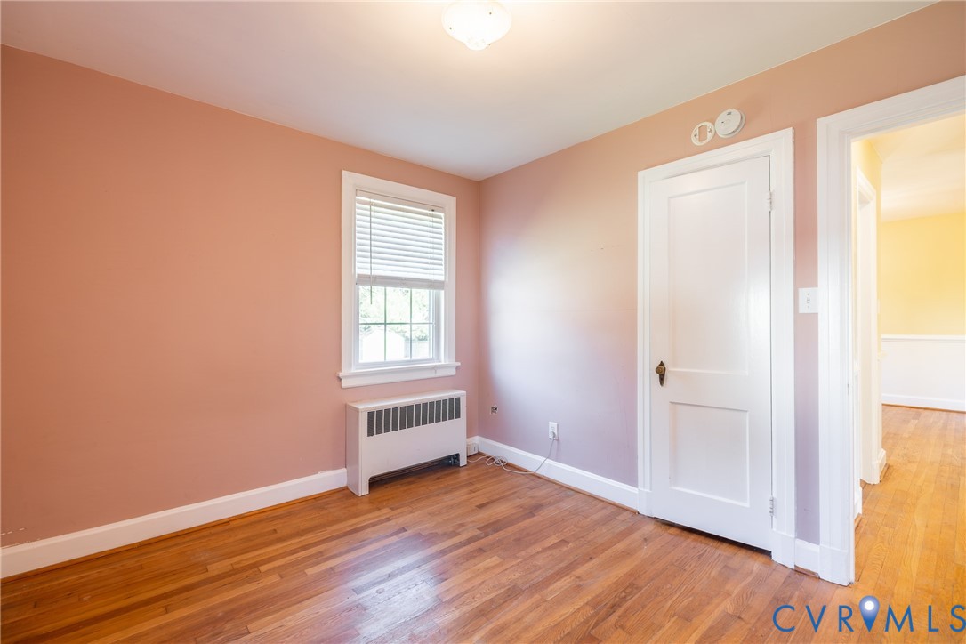 Room featuring wood-finish flooring, a single-pane window, a solid-panel door with brass hardware, and a metal radiator enclosure