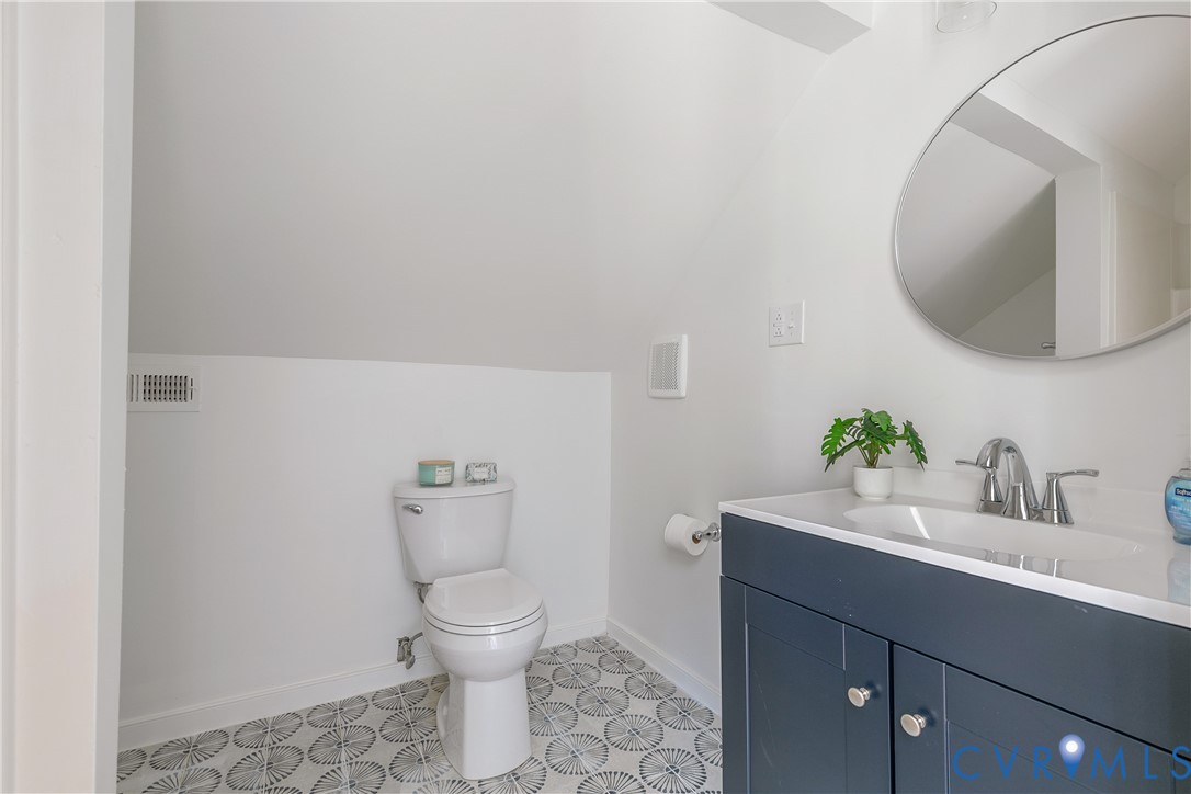 Bathroom featuring a white toilet, patterned floor tiles, a dark blue vanity with silver hardware, a white countertop with an integrated sink, and a circular mirror