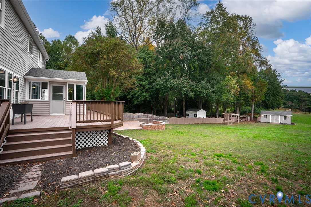 Raised deck featuring horizontal railings and lattice skirting, stone fire pit, paved patio area, and mature perimeter trees