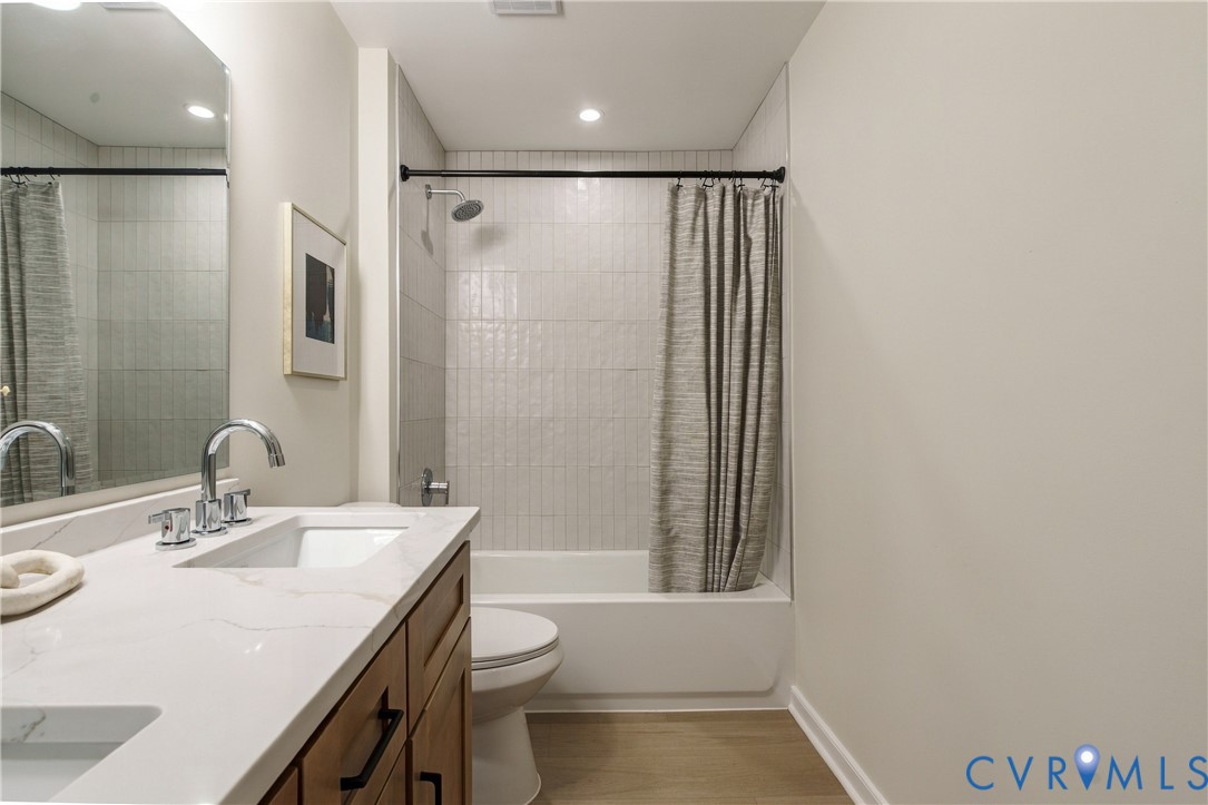 Bathroom featuring a vanity with white stone-finish countertop, dual under-mount sinks, chrome widespread faucets, wood-finish cabinetry, and a tub-shower combination with floor-to-ceiling tile surround