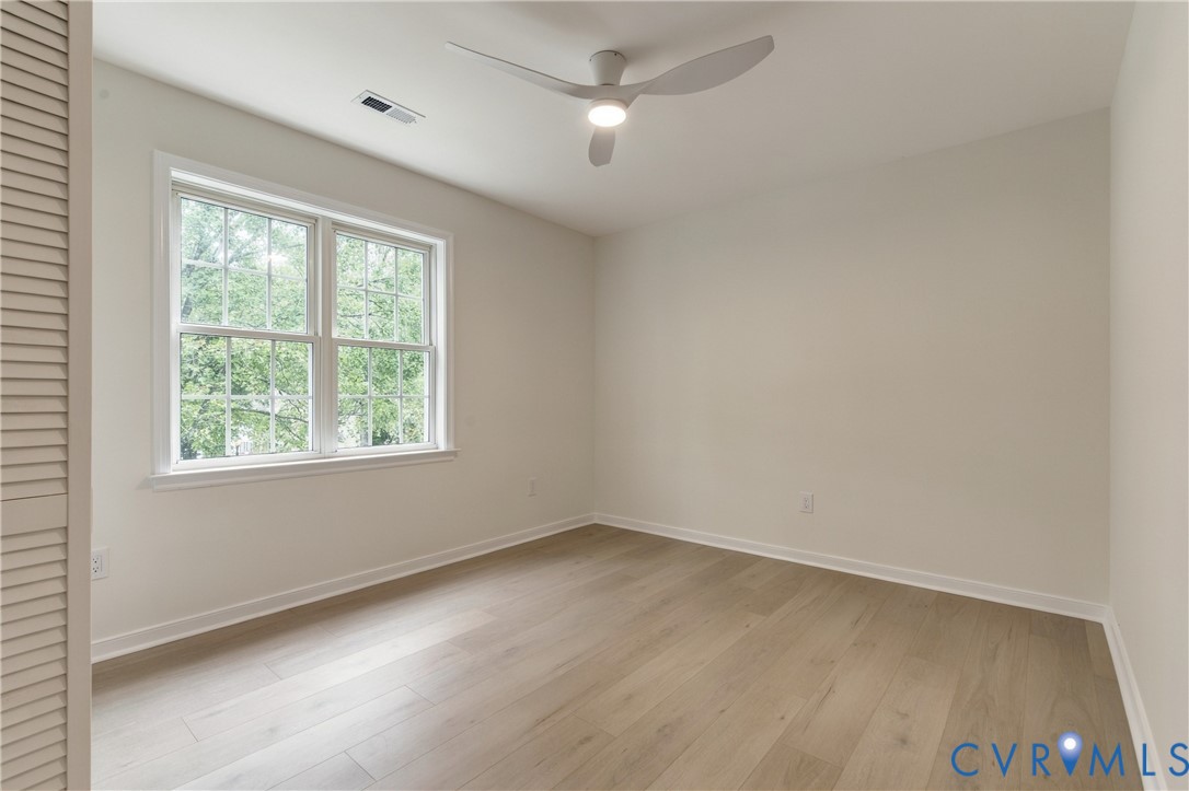Neutral-toned interior featuring wood-finish flooring, a multi-pane window, louvered closet door, and a modern three-blade ceiling fan with integrated lighting