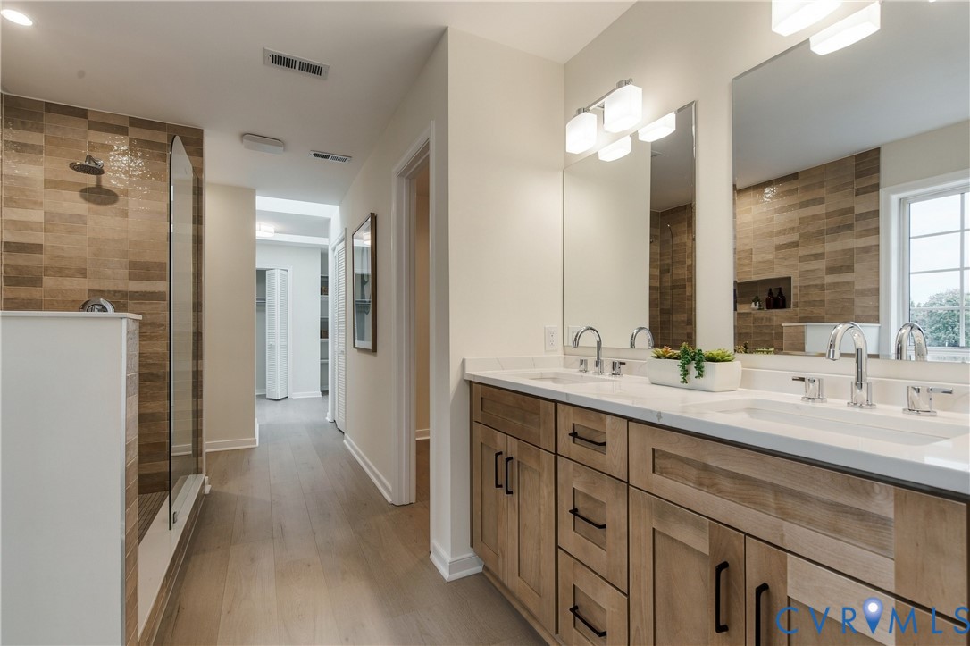 Bathroom featuring a dual-sink vanity with light wood cabinetry, white countertops, textured horizontal tile shower wall, and wood-finish flooring