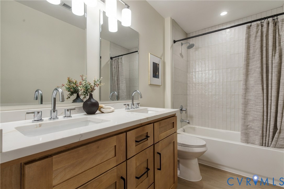 Dual-sink vanity with light-toned wood cabinetry, stone-finish countertops, wall-mounted light fixtures, and a tub-shower combination featuring vertically-laid tile surround