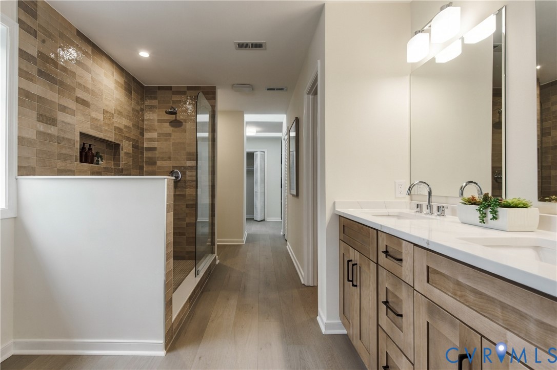 Double vanity featuring wood-finish cabinetry, white countertop with dual undermount sinks, wall-mounted light fixtures, and glass-enclosed walk-in shower with textured tile wall and built-in recessed niche