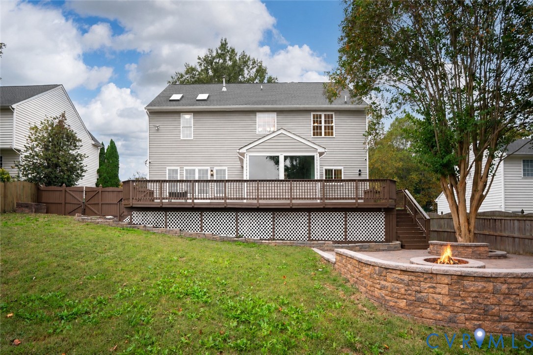 Rear exterior featuring a tiered wooden deck with lattice skirting, paver patio, stone-finish circular fire pit, and privacy fencing